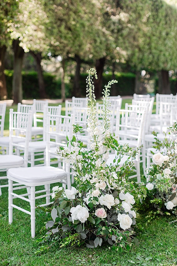 Ceremony aisle decor with outdoor wedding ceremony chairs beside a low white rose and eucalyptus arrangement on a grassy garden lawn