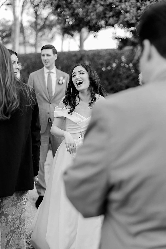 Wedding candid moment of a bride laughing candid in an off the shoulder gown with drop earrings, greeting guests in a garden setting
