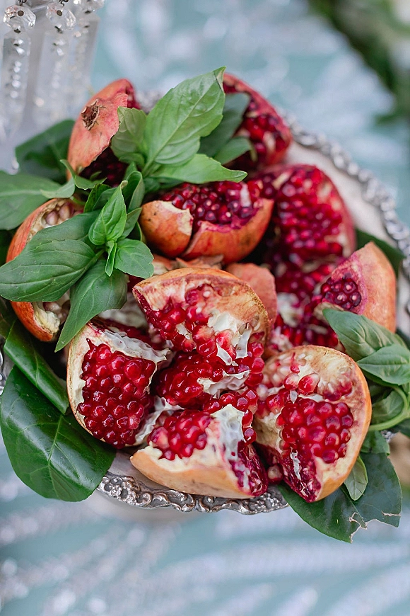 Wedding fruit platter with pomegranate fruit platter, basil leaves and greenery on a silver tray over light blue fabric with crystal beads