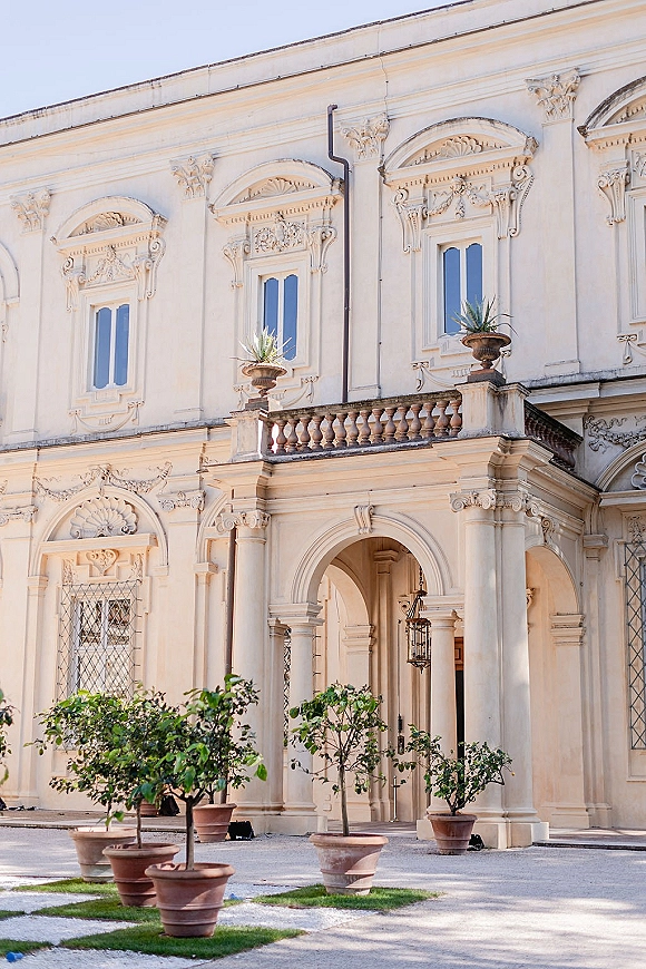 Wedding venue exterior with a historic arched doorway, columns, and hanging lantern, framed by terracotta planters in a stone courtyard