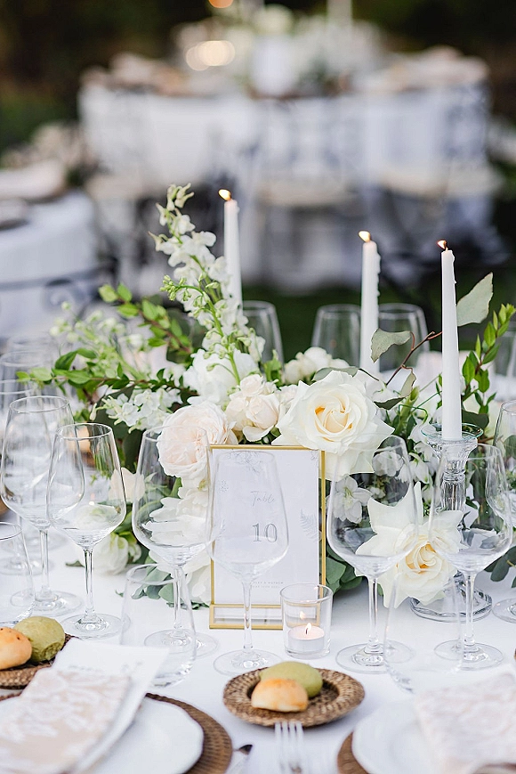 Reception tablescape with a white floral centerpiece and greenery garland, taper candles, and gold table number on an outdoor lawn under trees