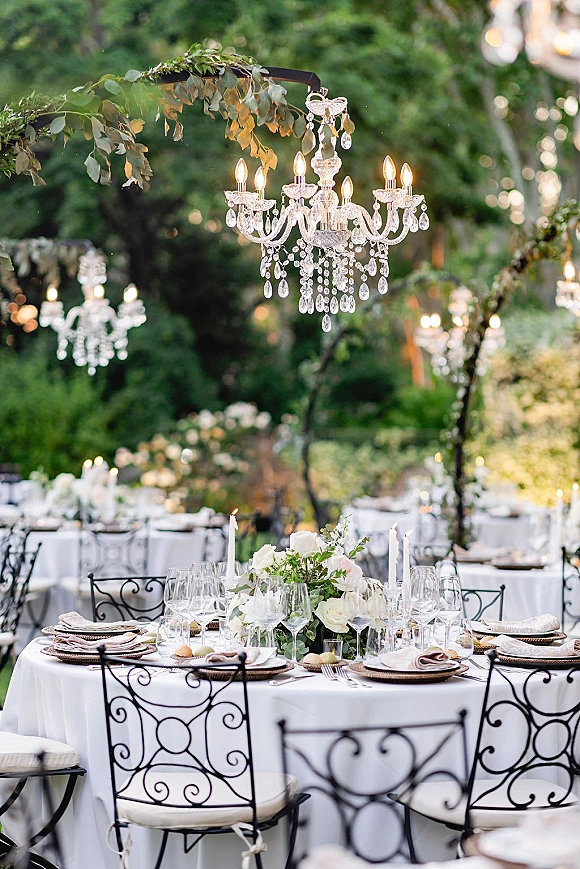 Reception tablescape with crystal chandeliers and hanging greenery, white linens, taper candles, and floral centerpieces in a garden setting