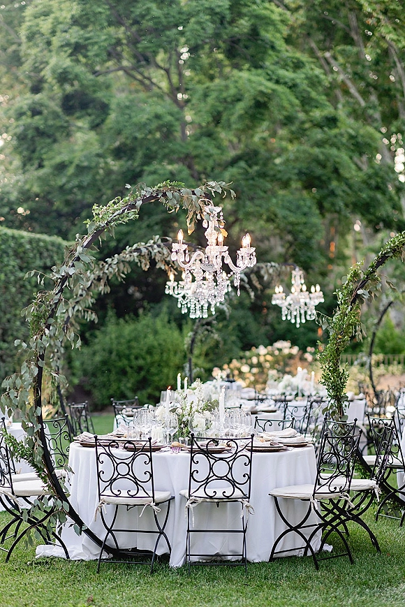 Outdoor reception tablescape with round white-linen tables, white floral centerpieces, taper candles, and crystal chandeliers on a garden lawn