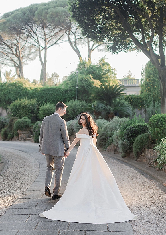 Couple portrait of bride and groom walking away holding hands, bride looking back in off-shoulder gown with long train on sunlit garden path