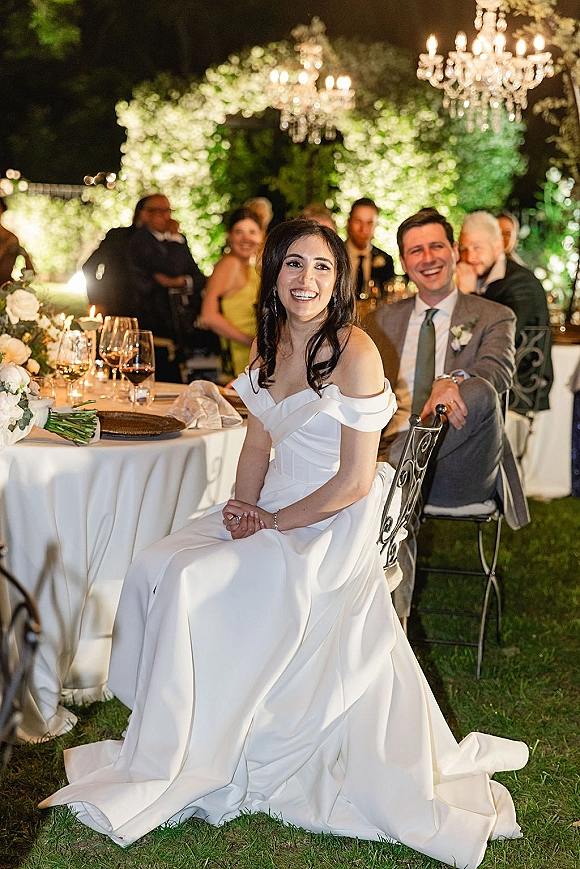 Reception moment as bride in an off the shoulder wedding dress laughs with groom at sweetheart table under chandeliers in a garden setting