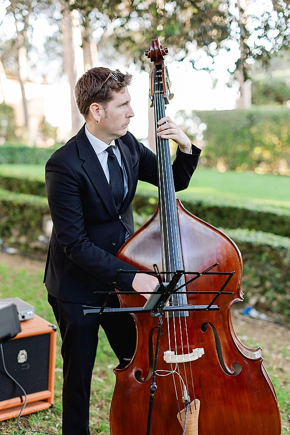 Wedding musician in sunglasses playing upright bass with sheet music and microphone, set on a daylight lawn with trees and hedges