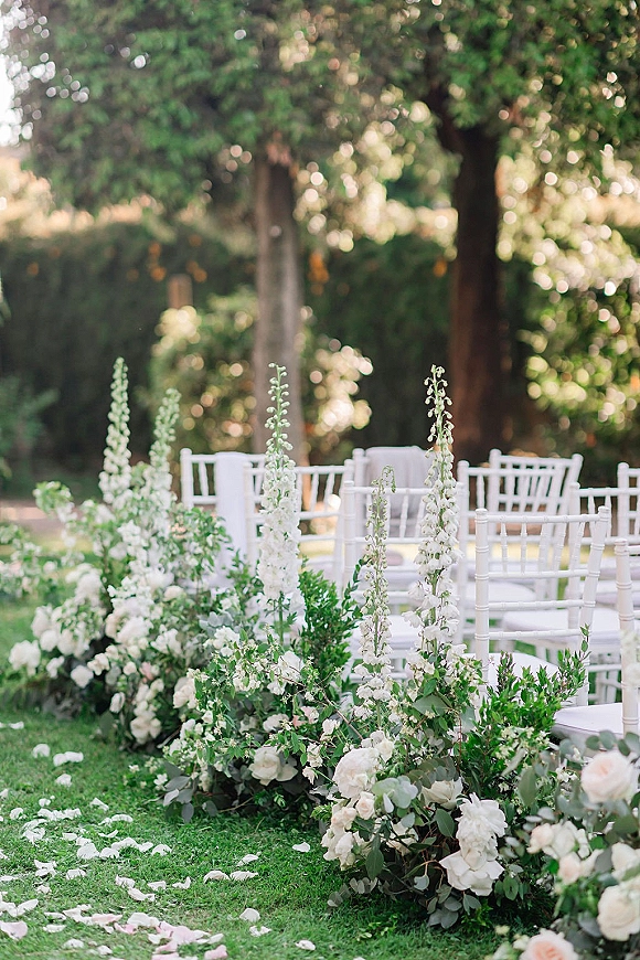 Ceremony aisle decor with outdoor ceremony aisle flowers, white rose and delphinium meadow arrangements and petal scatter on a garden lawn
