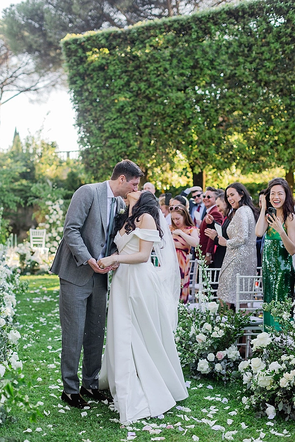 Ceremony kiss as bride and groom embrace on a petal-strewn aisle, veil flowing, guests filming amid white rose flowers and greenery on lawn