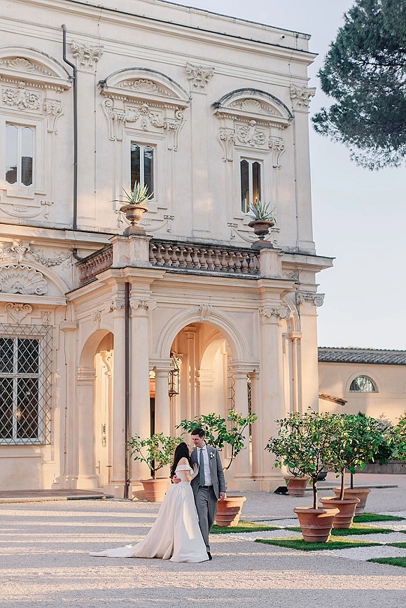 Couple portrait of bride and groom embrace in an off-the-shoulder gown and gray suit before an ornate villa archway and courtyard citrus trees