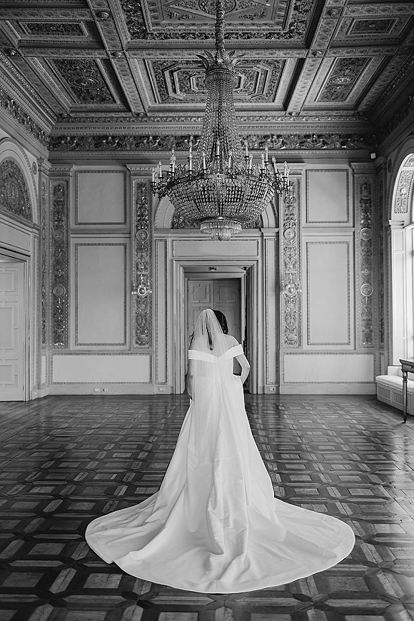 Bridal portrait of a bride from behind with a cathedral length veil and dramatic wedding dress train in an ornate ballroom under a chandelier