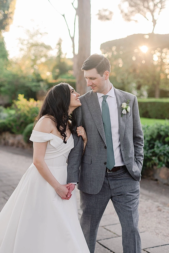 Couple portrait of bride and groom holding hands on a garden path, bride looking up at groom in off-the-shoulder dress with sun flare