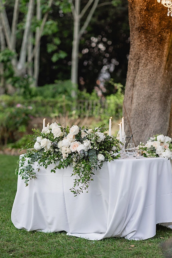 Reception tablescape with white floral centerpiece and greenery garland, taper candles and glassware on a white tablecloth under a crystal chandelier outdoors