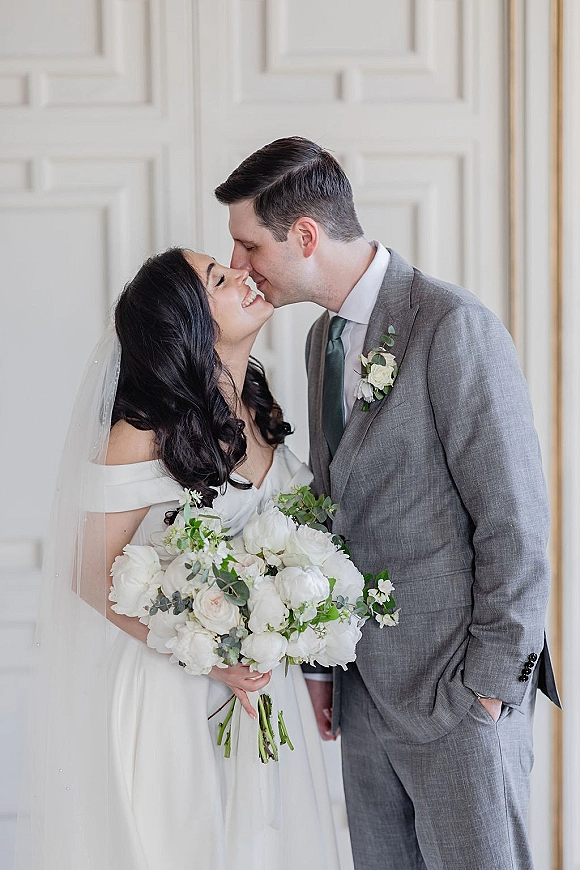 Wedding couple portrait of bride and groom kiss, bride in veil holding white rose bouquet beside groom in gray suit with green tie against white paneled wall
