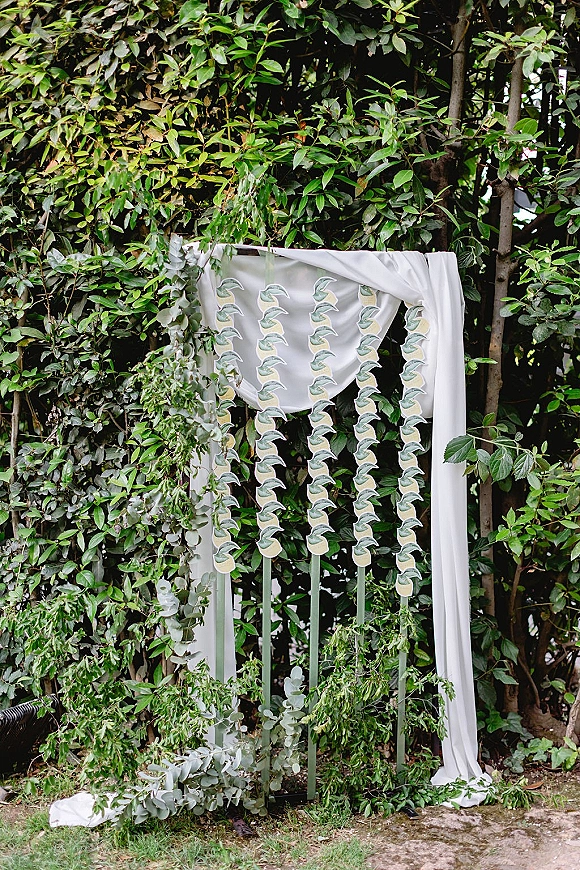 Ceremony backdrop with white drapery and hanging greenery garlands, set on a grass lawn with garden trees and shrubs behind