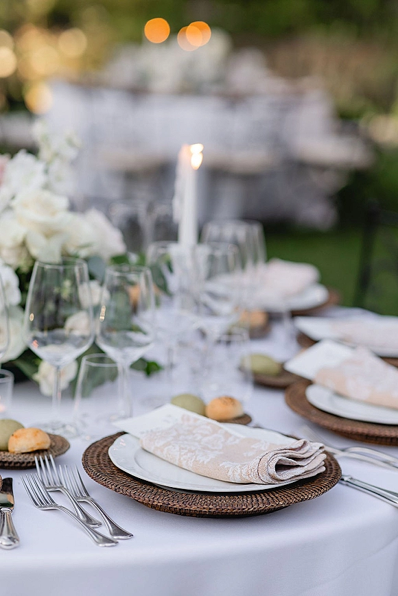 Reception tablescape with rattan chargers, white plates, linen napkins, taper candle, and floral centerpiece under garden bistro lights