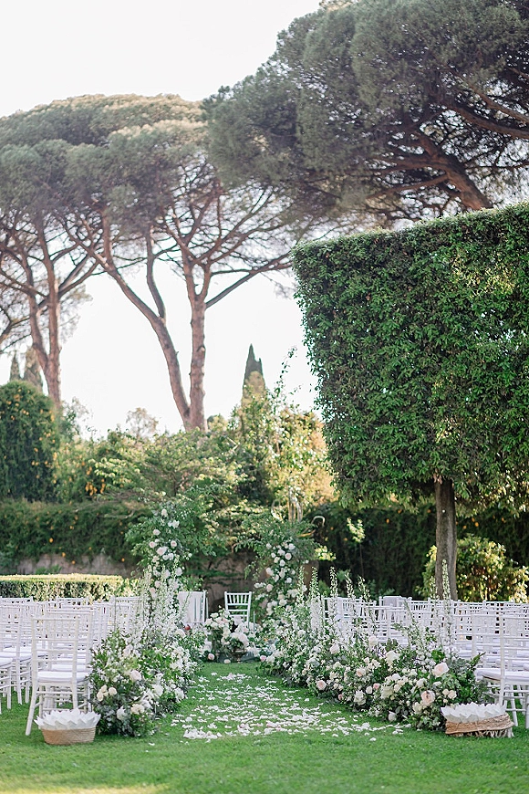 Ceremony setup for an outdoor wedding ceremony with white chairs, white rose aisle flowers, petal runner, and floral arch on a garden lawn