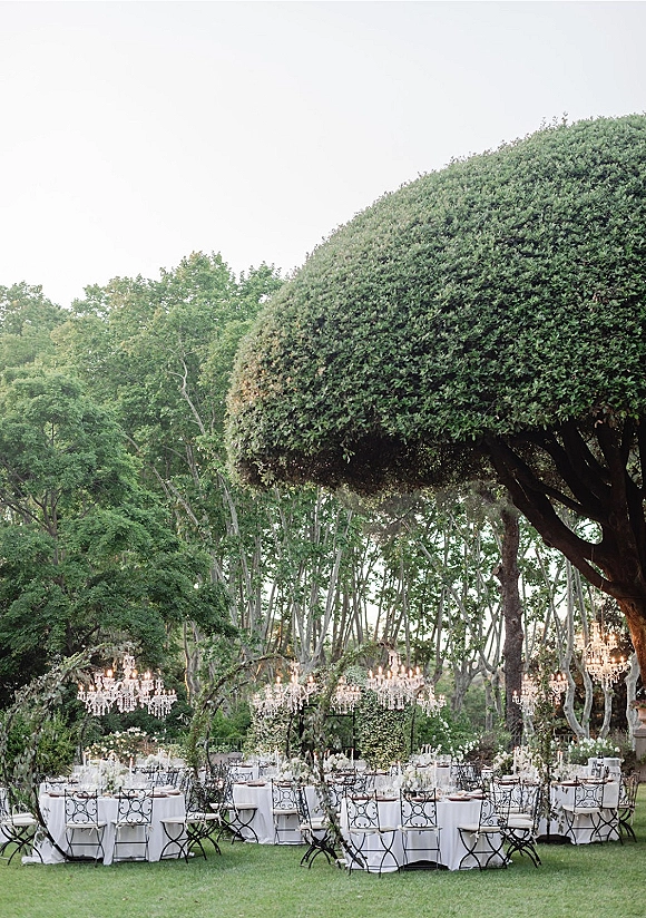 Outdoor reception decor with round tables in white linens, wrought iron chairs, taper candles, florals and crystal chandeliers under tree canopy