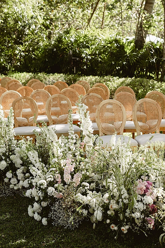 Ceremony seating with cane back wedding chairs and white cushions lining an aisle of white and pink ground flowers on a sunlit garden lawn