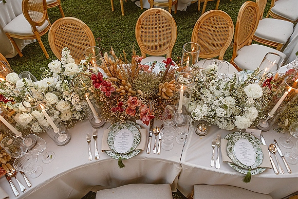 Reception tablescape with wedding head table decor, long floral centerpiece of roses and dried grasses, taper candles in hurricanes on a lawn