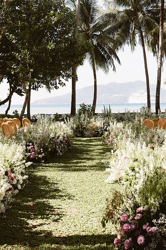 Ceremony aisle decor with pastel ground floral arrangements lining grass between rattan chairs, facing palm trees and ocean view beyond