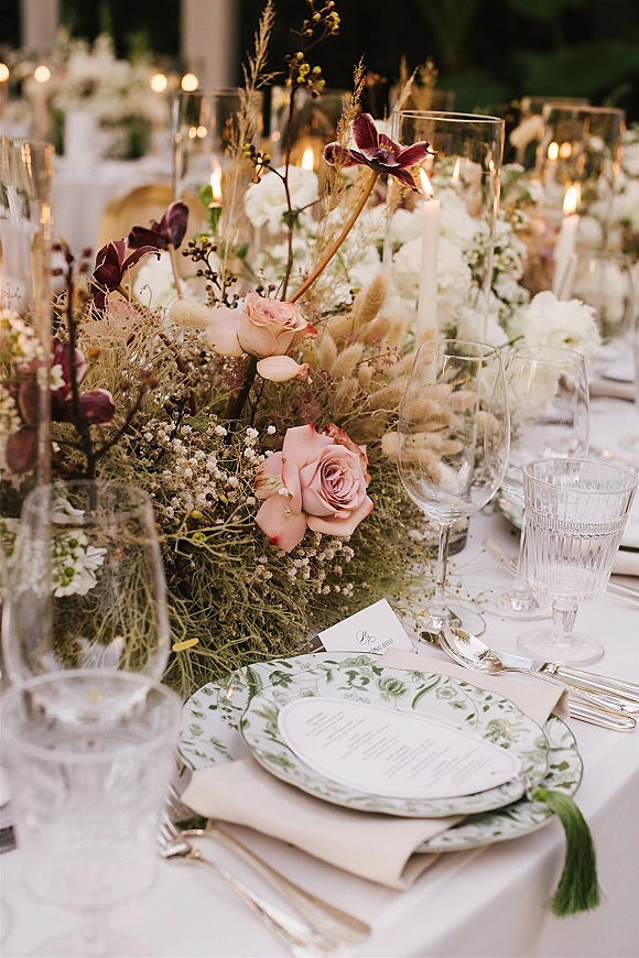 Reception tablescape with wedding table centerpiece of blush roses and dark purple orchids, taper candles, bud vases, and printed menus on plates