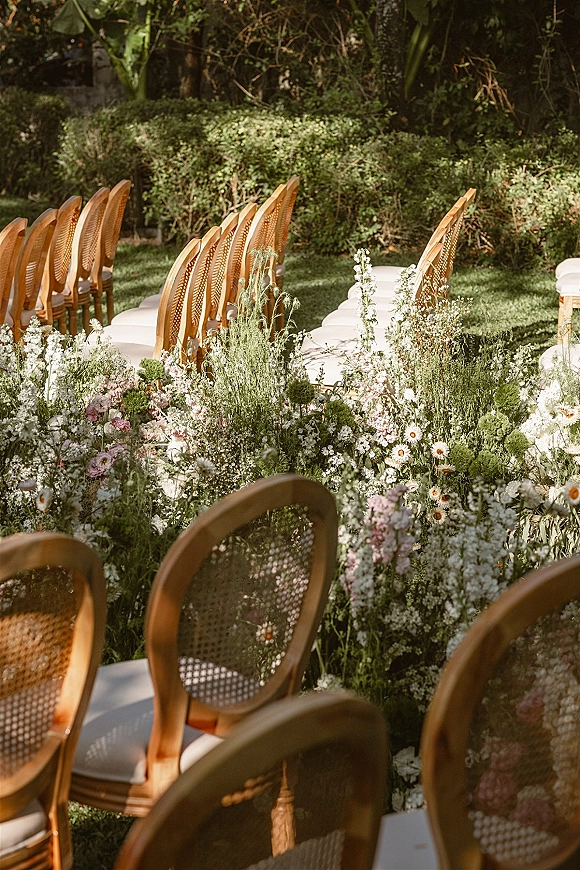 Ceremony aisle decor with outdoor wedding aisle flowers, cane-back chairs and white cushions lining a runner edged with wildflowers on a sunny lawn