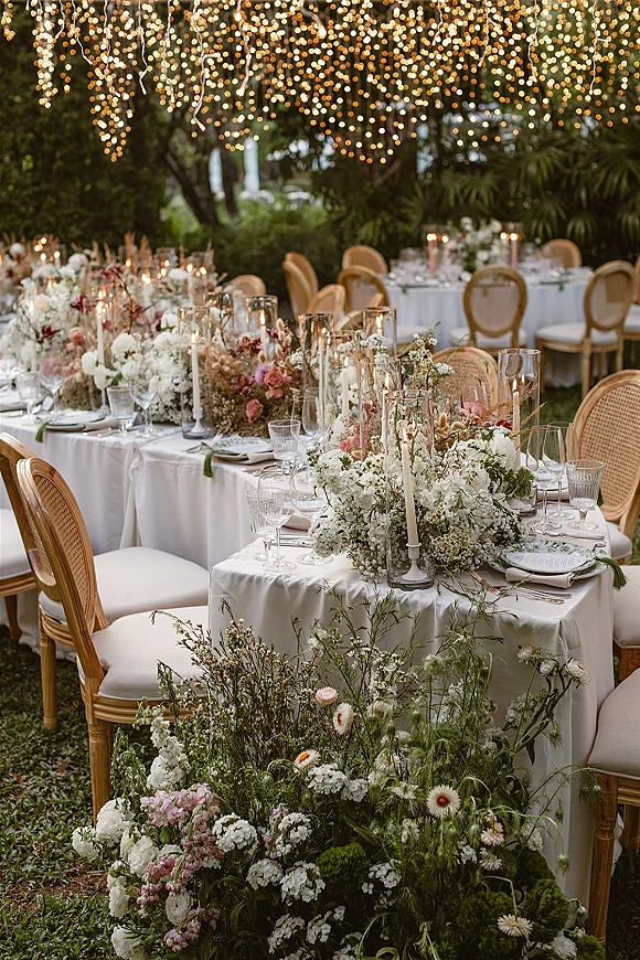Reception tablescape with white linens, floral centerpieces, taper candles in hurricanes, and rattan chairs under string lights in a garden setting