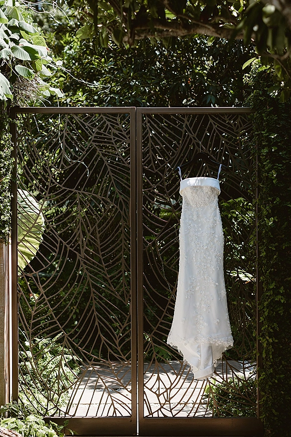 Wedding dress on a hanger, lace wedding dress with spaghetti straps, hanging from a metal garden gate amid lush sunlit greenery