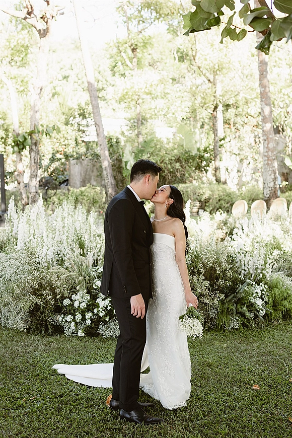 Wedding kiss portrait of bride and groom kissing, her strapless lace gown and white bouquet against a lush garden lawn with white flowers