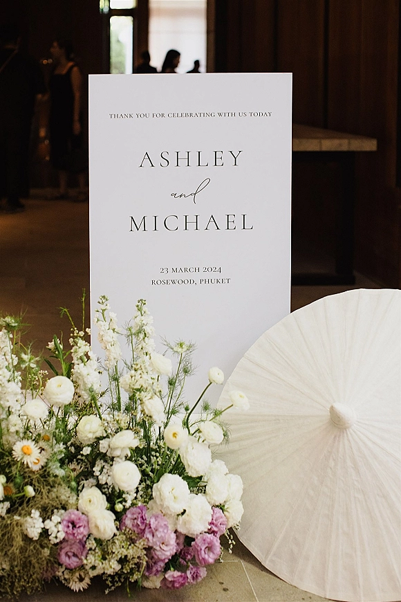Wedding welcome sign with flowers on a white board, framed by purple and white blooms and greenery at an indoor entrance with wood wall