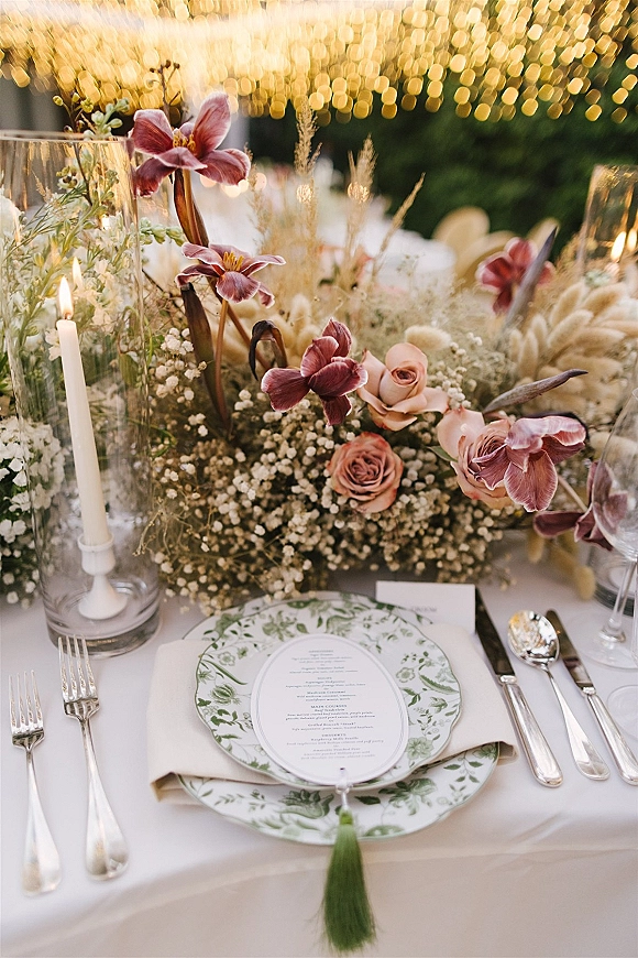 Reception tablescape with wedding place setting, rose and orchid centerpiece, taper candles in glass holders, and string lights in greenery backdrop