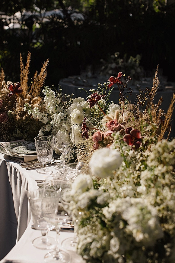 Reception tablescape with wedding table centerpiece of white, blush and burgundy blooms, crystal stemware, set on a sunlit garden table
