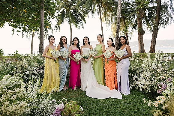 Bridesmaid group portrait with bride with bridesmaids holding bouquets in colorful dresses, posed on a lawn with palm trees and ocean behind