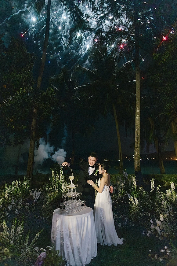 Champagne tower toast as bride and groom pour bubbly into stacked coupe glasses on a white table, with fireworks and palm trees at night