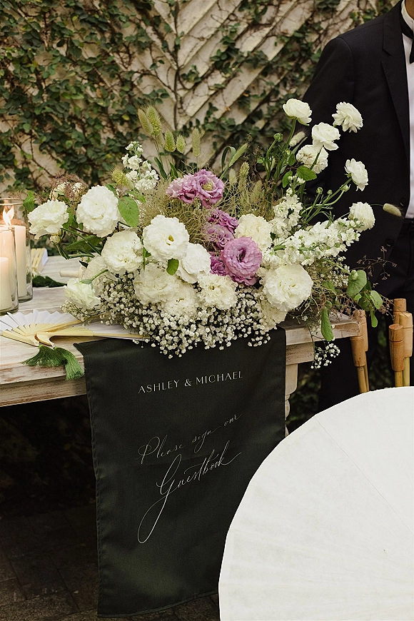 Wedding guestbook table with wedding guestbook sign, white and lavender floral centerpiece, candles, fans and champagne bottles against an ivy wall