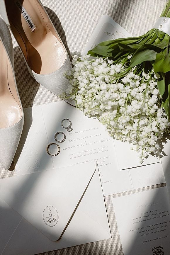 Invitation suite with minimal wedding invitations arranged on linen fabric with envelope, rings, bridal shoes, and lily of the valley bouquet