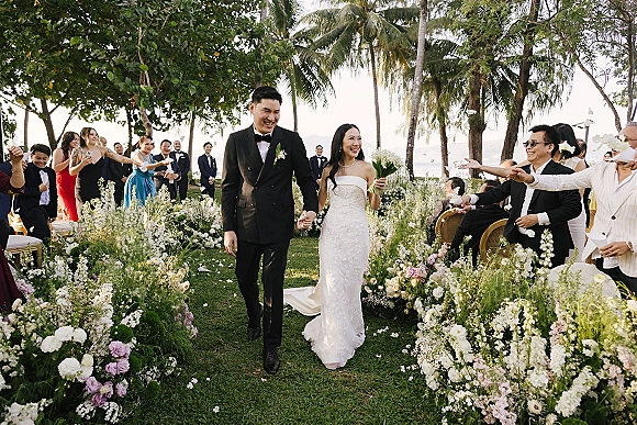Wedding recessional as bride and groom walk hand in hand down the aisle, guests tossing petals on a palm-lined oceanfront lawn