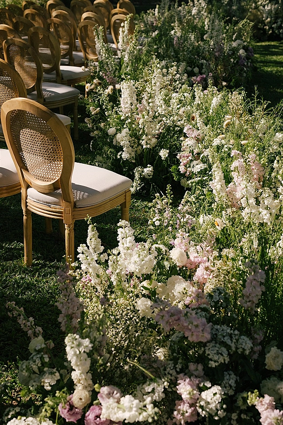 Ceremony aisle flowers in a wildflower meadow style with pastel pink and white blooms and greenery beside cane-back chairs on a sunny lawn