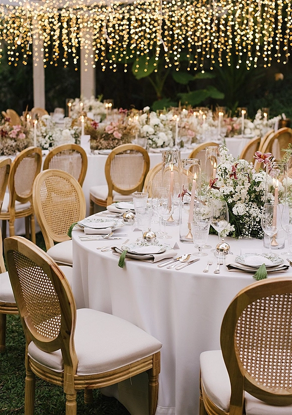 Reception tablescape with round tables, white tablecloths, floral centerpieces, taper candles in hurricanes, and string lights in a garden pergola setting