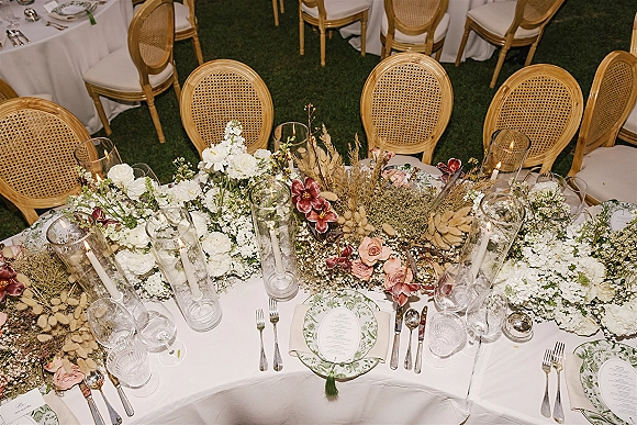 Reception tablescape with head table decor, white hydrangea and rose garland, taper candles in glass hurricanes, on a grass lawn