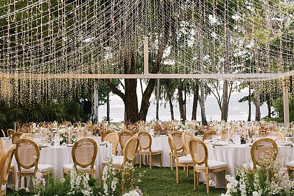 Outdoor reception decor with string light canopy over round tables, white linens, rattan chairs, floral centerpieces and candles by the water view