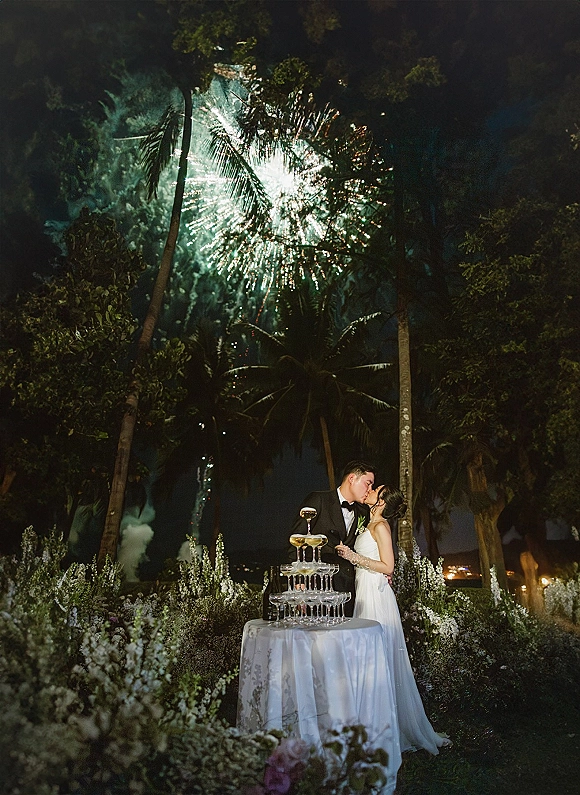 Wedding kiss as newlyweds embrace by a champagne tower accent, with fireworks lighting the night sky and palm trees behind them