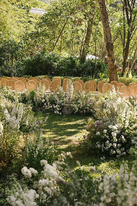 Ceremony aisle design with a floral ground border of white and blush blooms and greenery, lined with wicker chairs in a sunlit garden lawn