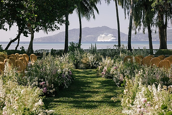 Ceremony aisle decor with meadow-style florals lining a grass aisle, flanked by cane back chairs, with palm trees and ocean view beyond