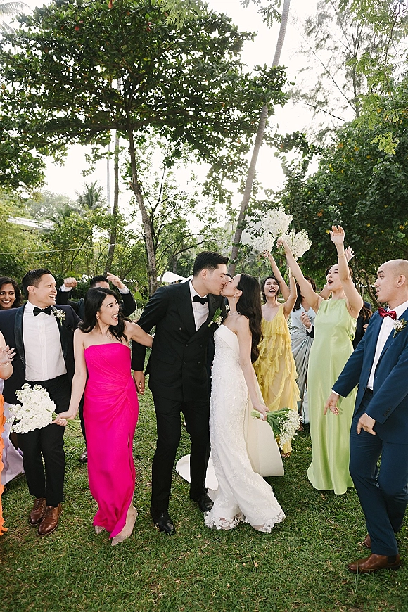 Wedding kiss as bride in veil and groom in tux share a kiss while bridesmaids and groomsmen cheer on a garden lawn