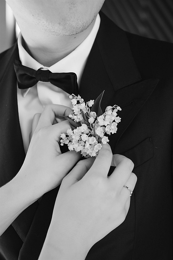 Groom boutonniere with lily of the valley boutonniere pinned on a black tuxedo lapel with bow tie against a dark indoor background
