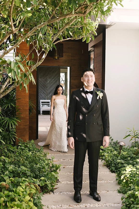 Wedding first look as bride approaches groom from behind on a stone path, lace train flowing beside a wood wall and garden greenery