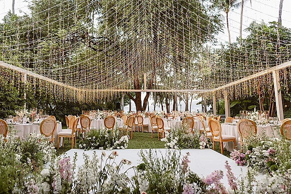 Reception tablescape with outdoor wedding reception round tables, white linens, rattan chairs, florals and candles under string lights by the ocean