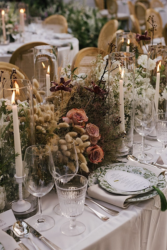 Reception tablescape with wedding table centerpiece of roses, greenery and dried florals, taper candles in hurricanes on white linens