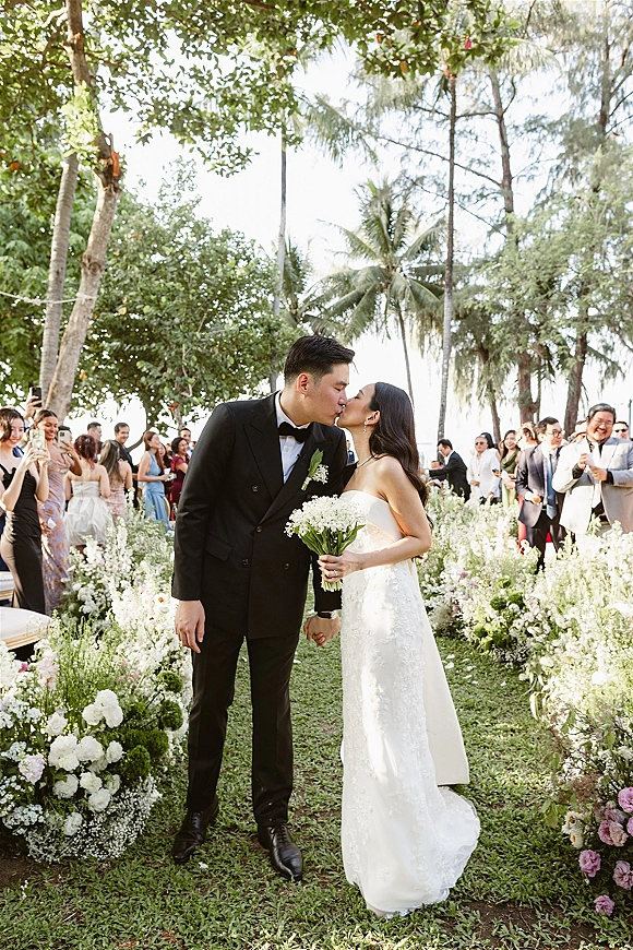 Wedding kiss as bride in strapless lace dress holds a bouquet, kissing groom in tuxedo beside flower-lined aisle in palm garden